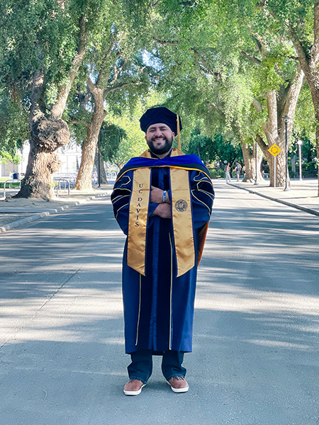 SF BUILD Dr. Juan Castillo stands on the street in his graduation regalia, in front of some trees