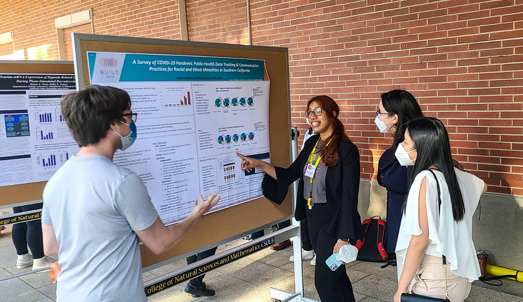 Outdoor photo of 4 people (CSULB BUILD students) looking at a poster at the Undergraduate Research Poster Session.