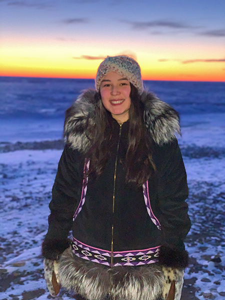 Samantha Wade stands in front of a sunset in Alaska wearing a thick coat. 