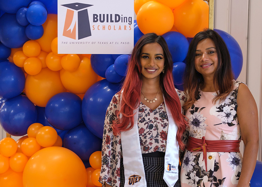 Aparna Mangadu & her mother Thenral Mangadu, Ph.D. stand together warmly embracing each other at a graduation event. There are blue and orange balloons and a sign with text “BUILDing Scholars The University of Texas at El Paso” behind them.